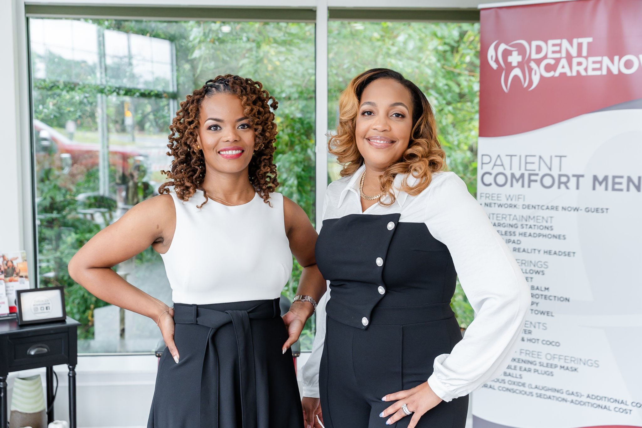 Two team members smiling in a Dentcare Now clinic reception area with patient comfort amenities displayed on signage behind them